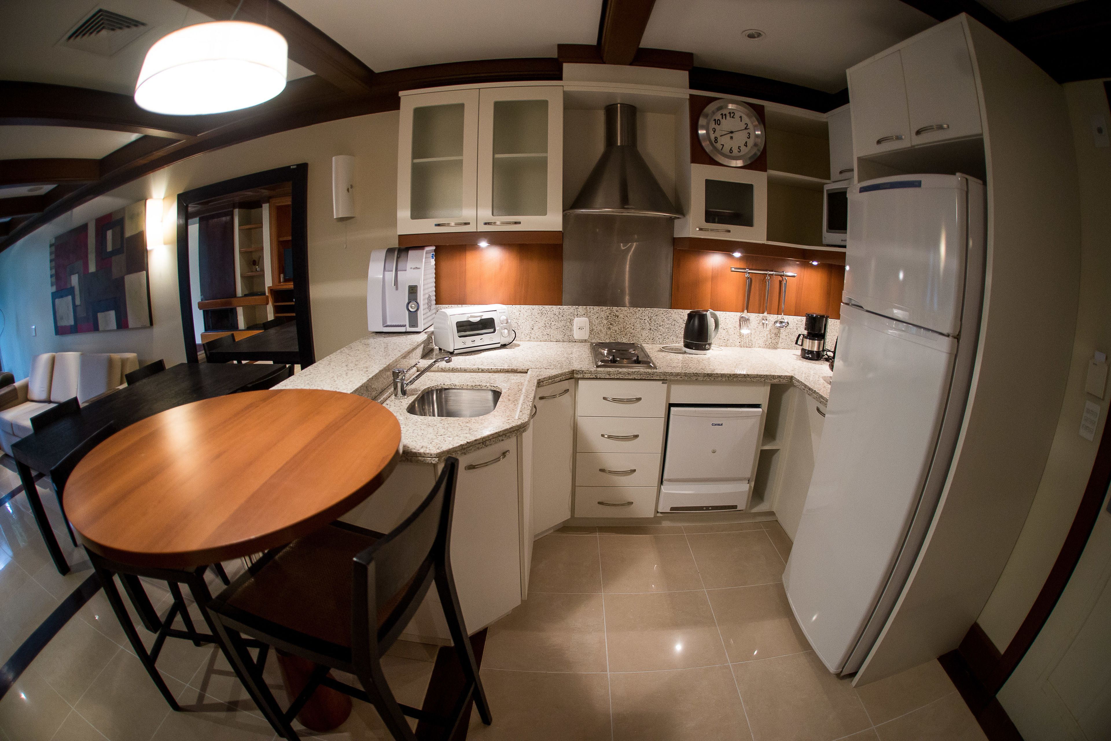 Cocina compacta con mesa de comedor de madera, armarios blancos, extractor de acero inoxidable y refrigerador blanco.