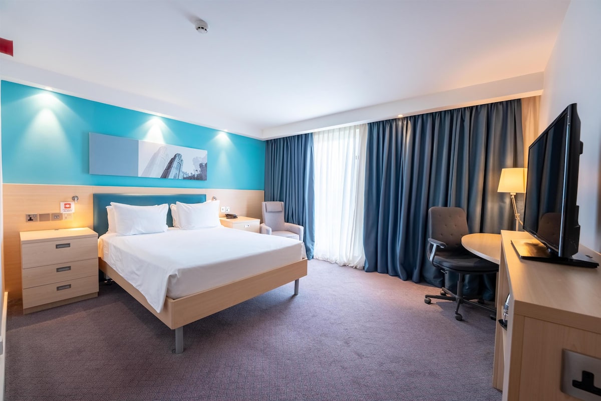 A hotel room with a bed, bedside tables, a desk with a chair, a TV, and a large window with curtains.
