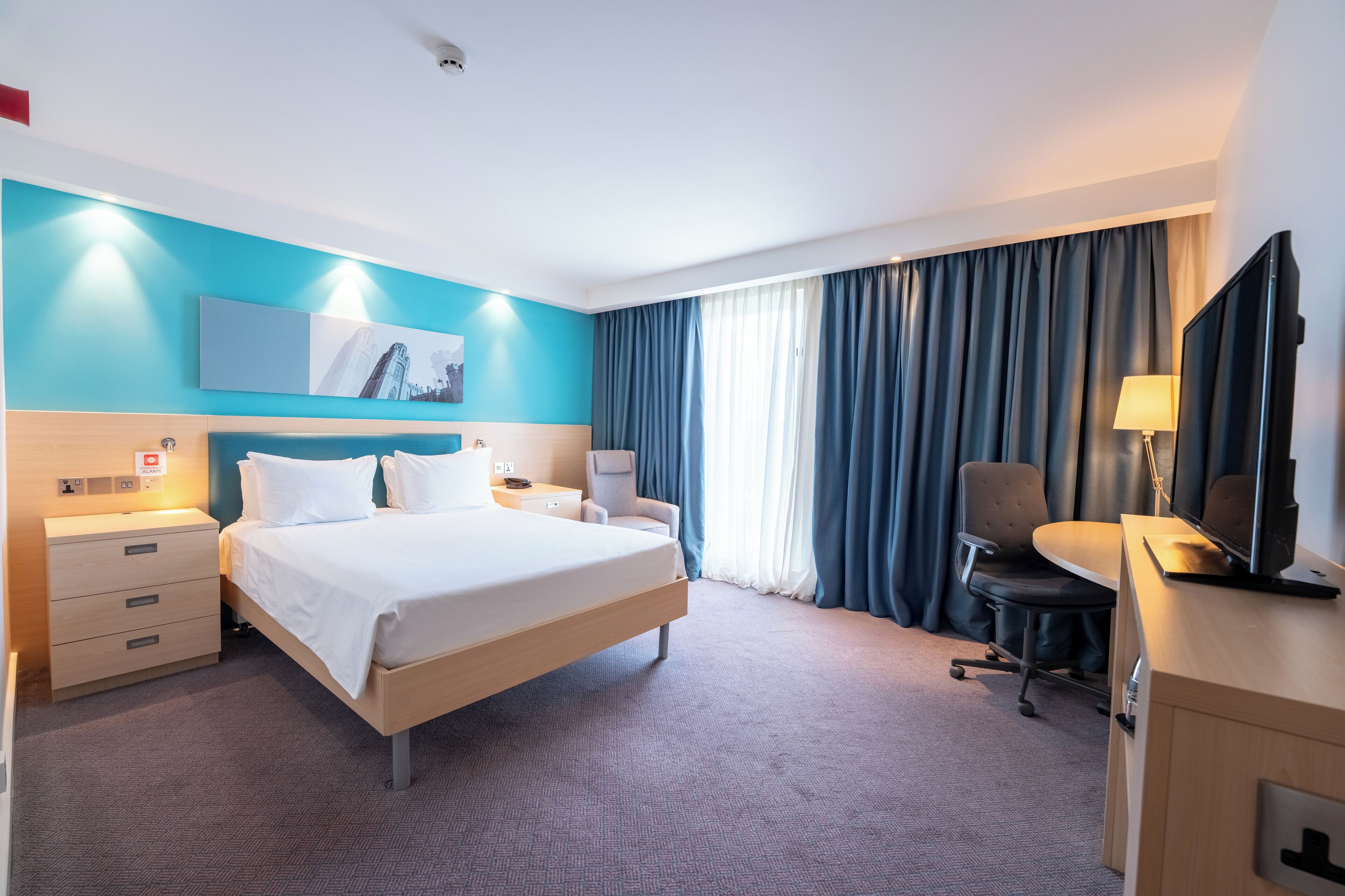 A hotel room with a bed, a desk with a chair, a television, and a large window with curtains.
