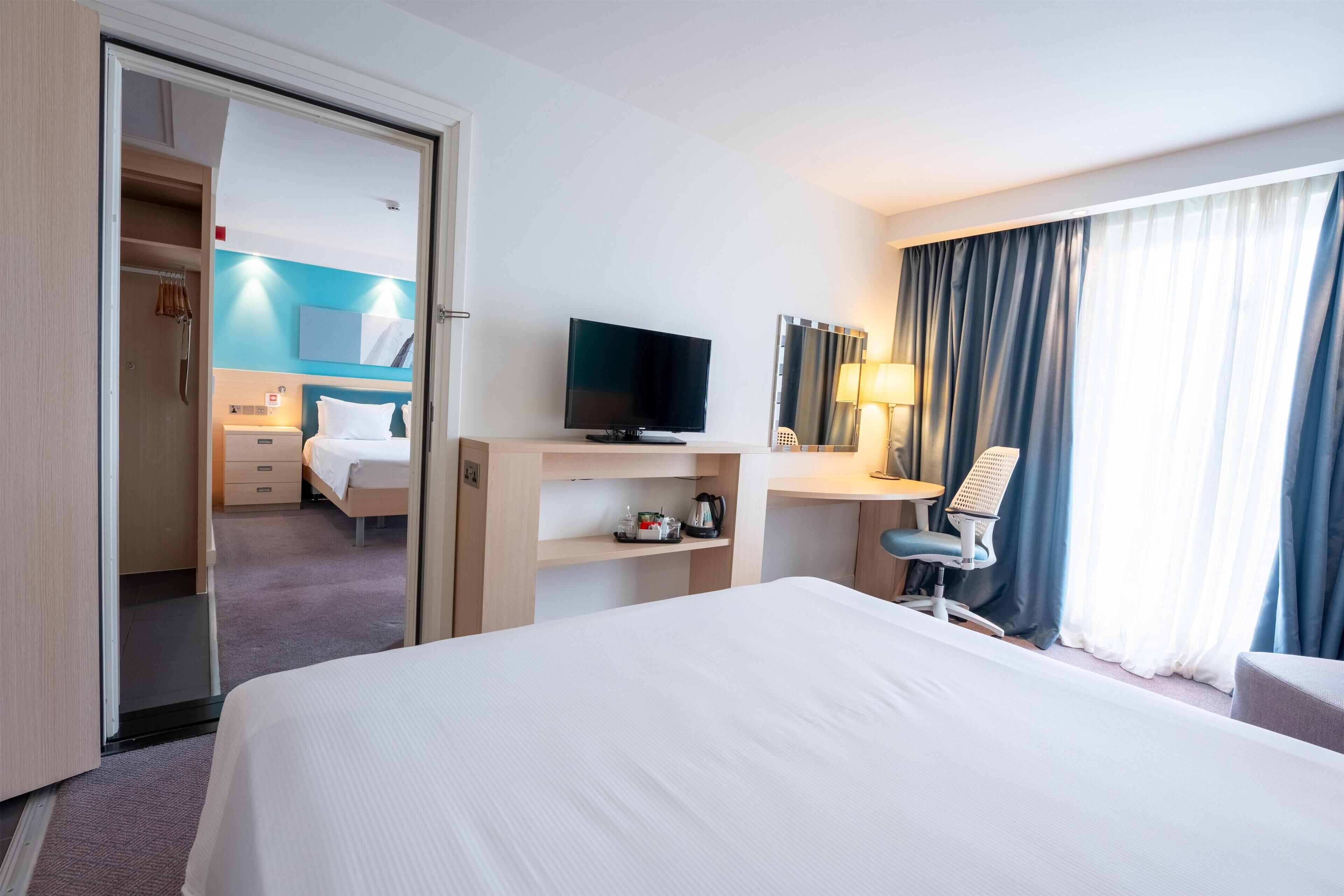 Une chambre d’hôtel comprenant un lit, un bureau, une télévision, une chaise, une lampe de bureau et une fenêtre avec des rideaux.