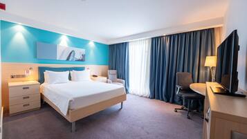 Een hotelkamer met een bed, een bureau met een computer, een stoel, een televisie en een raam met gordijnen.