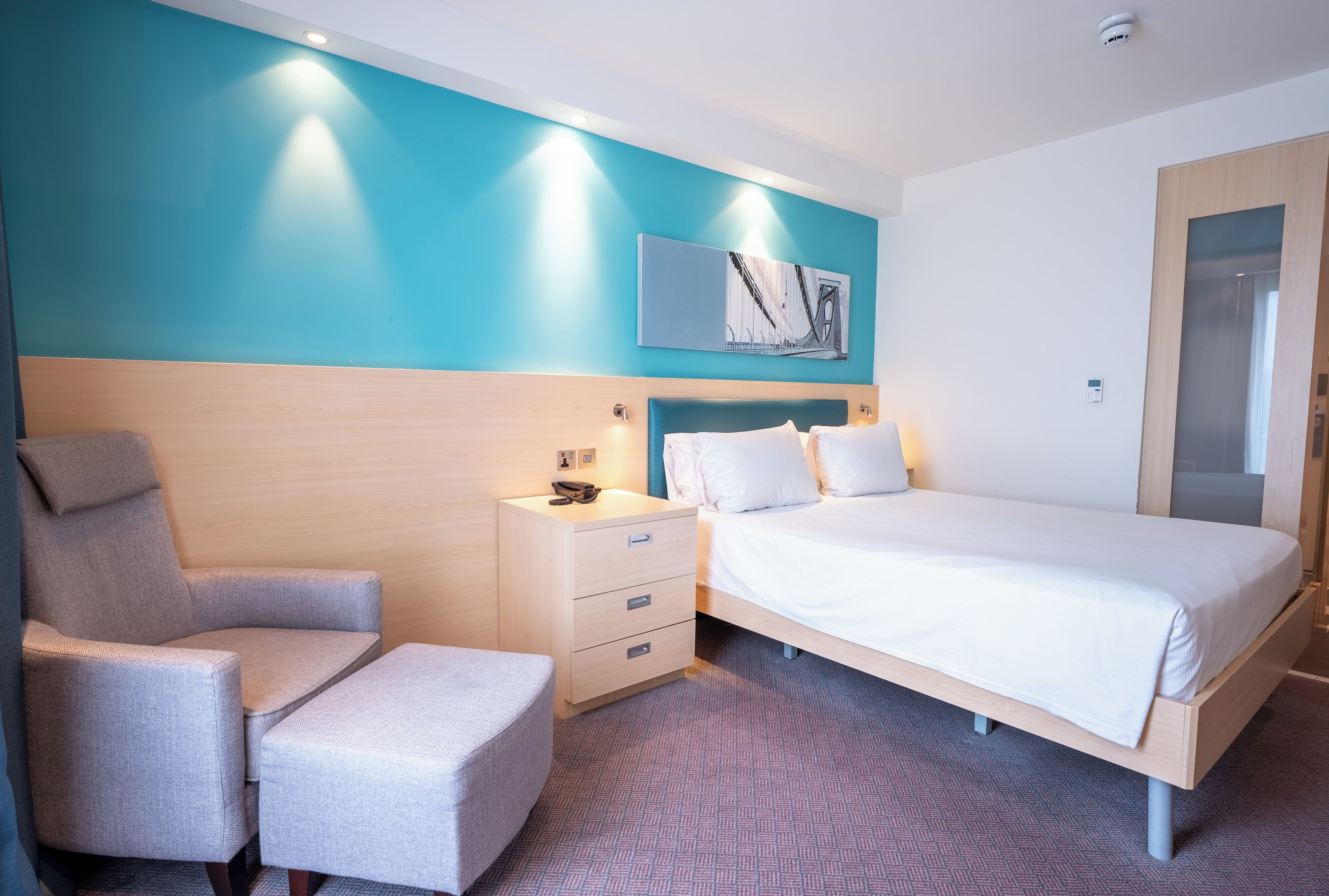 Een hotelkamer met een bed, een stoel, een nachtkastje en een kunstwerk aan de muur.