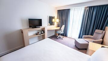 A hotel room with a large bed, a desk with a television, a chair, and a desk lamp.