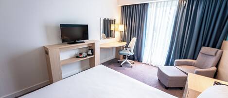 Habitación de hotel con una cama grande, un escritorio con televisor, una silla y una lámpara de escritorio.