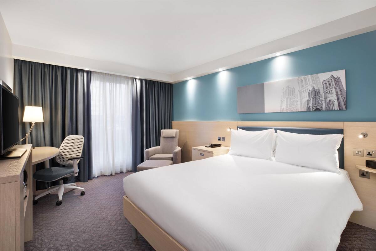 A hotel room with a large bed, a desk with a chair, a TV, and a painting on the wall.