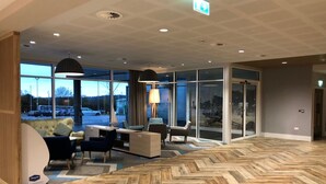 Um hall de hotel moderno com piso de madeira em padrão herringbone, área de estar com sofás e cadeiras, janelas amplas com vista para o estacionamento e um balcão de recepção.