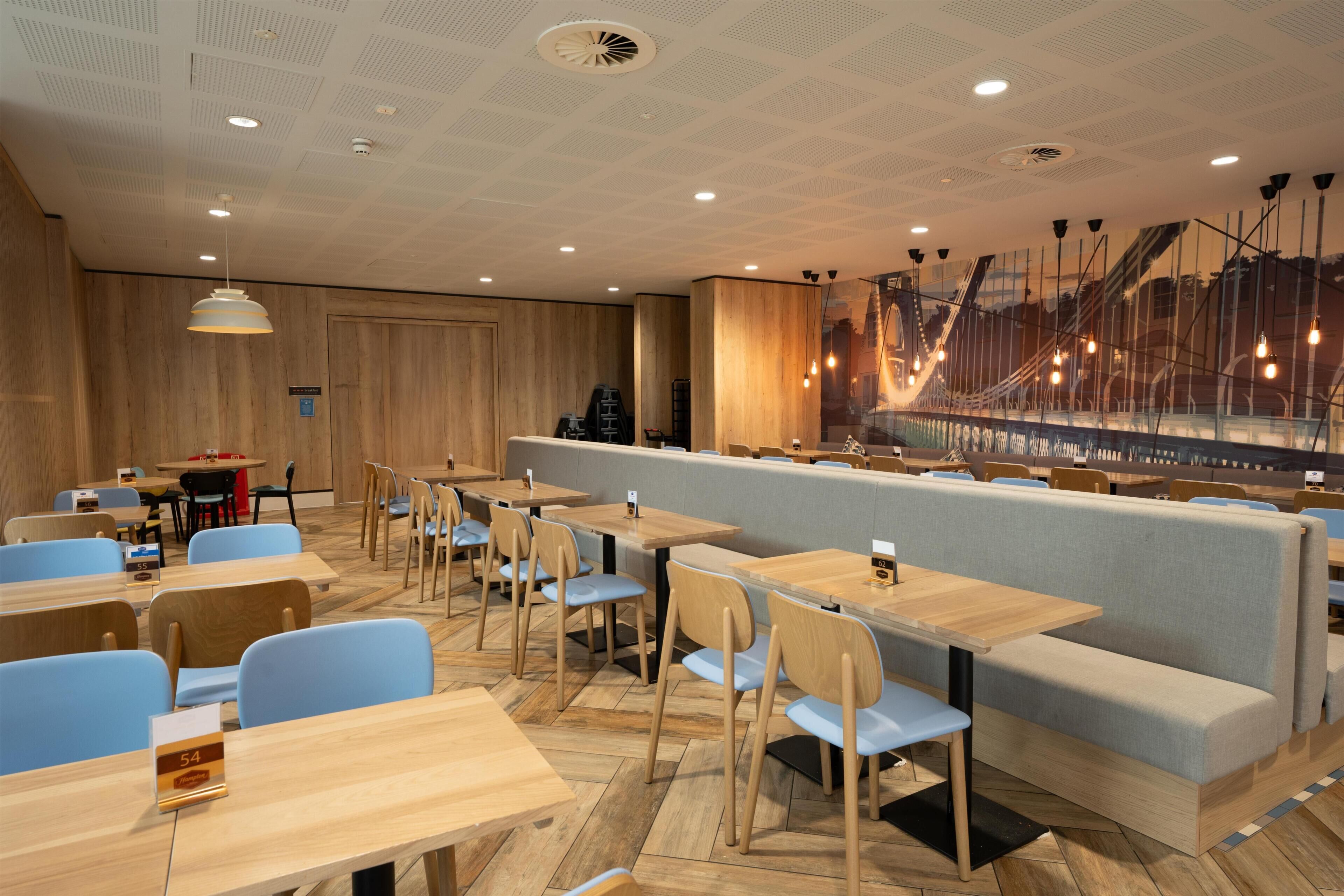 En modern restaurang med träbord och blå stolar, en sittgrupp med en vadderad bänk och ett dekorativt glasräcke.