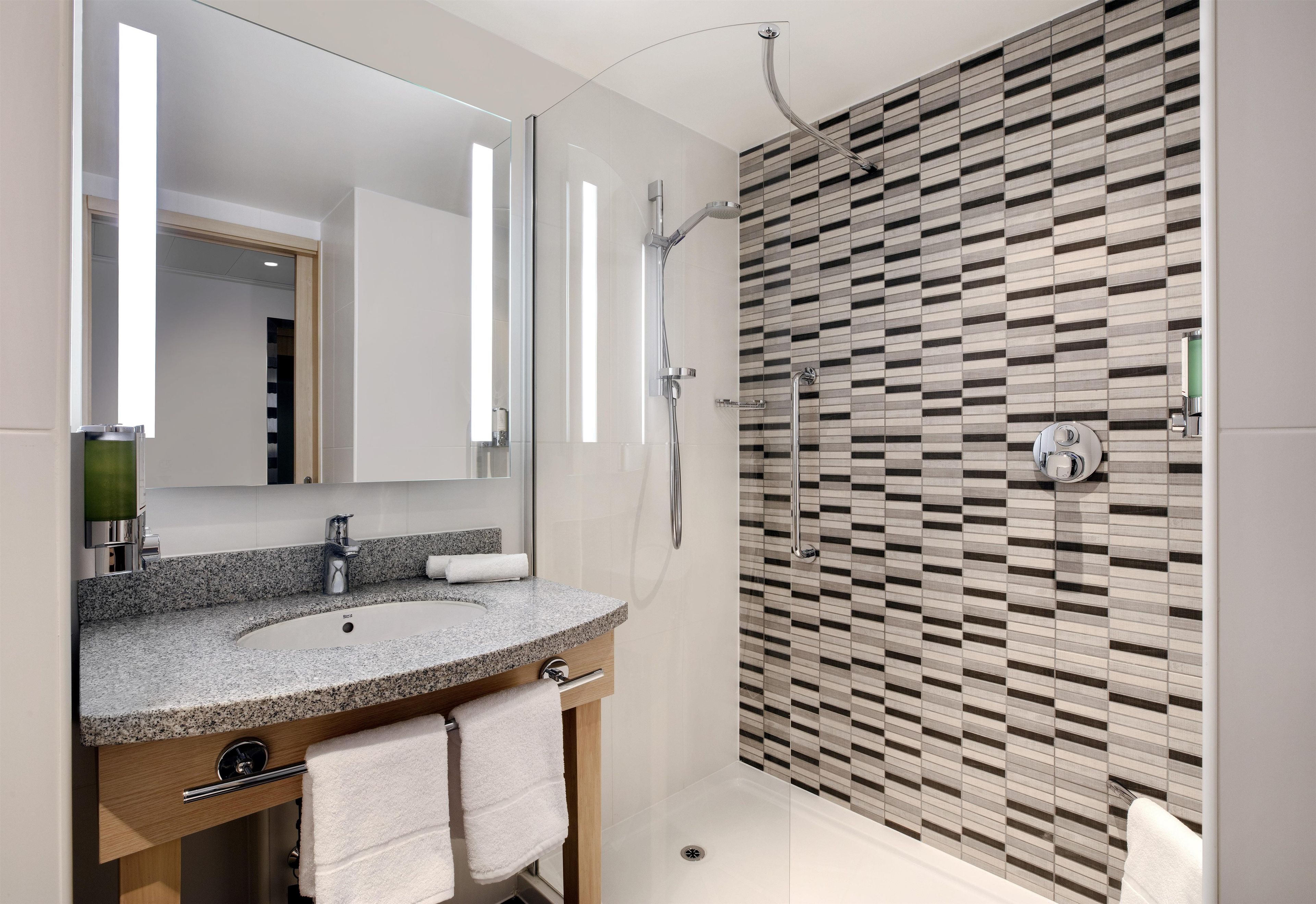 A modern bathroom with a granite countertop sink, a mirror, a shower with a glass door, and a patterned wall.