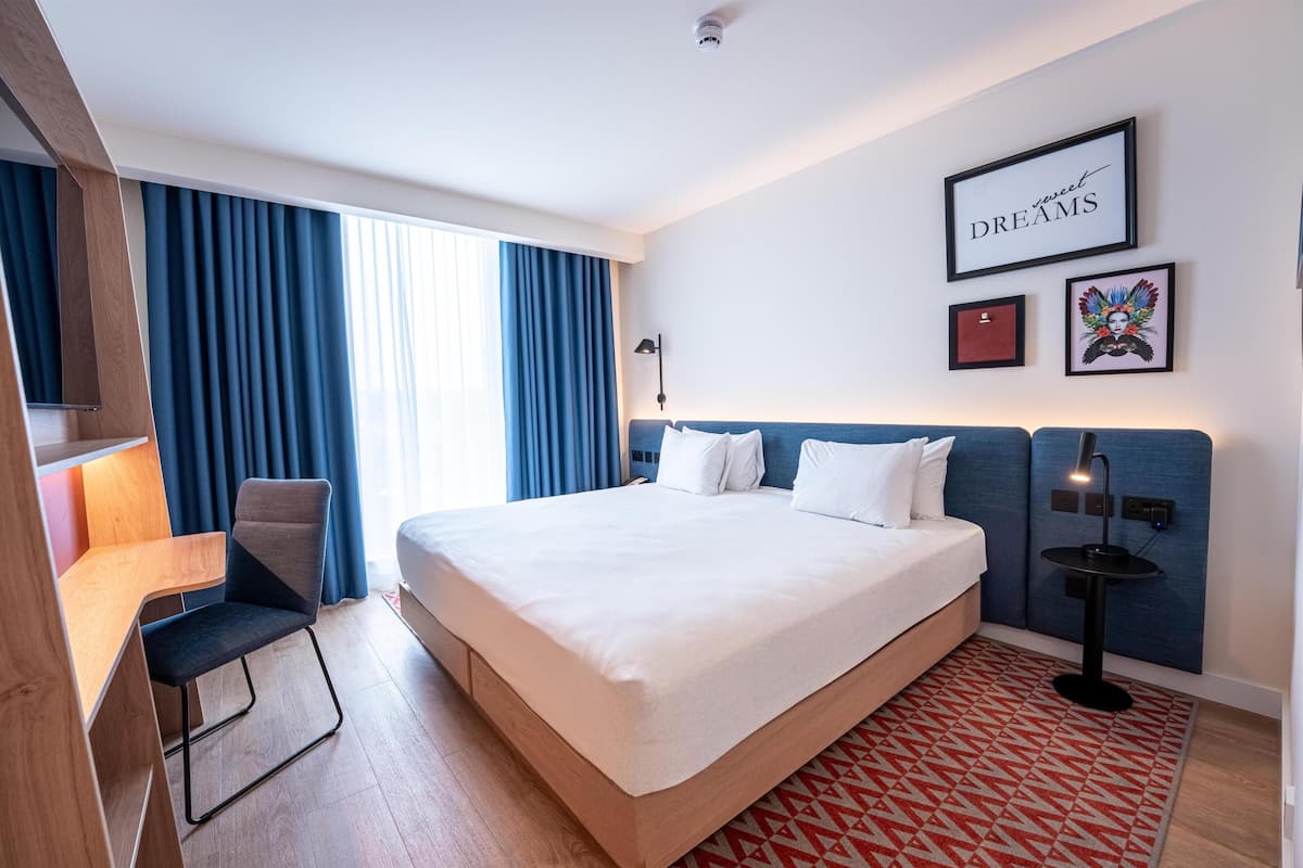 A hotel room with a large bed, a desk, a chair, a desk lamp, a wall-mounted TV, and a framed picture on the wall.