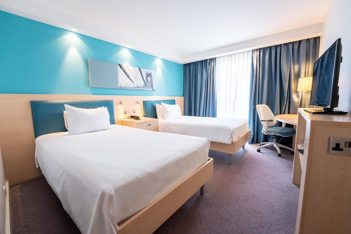 A hotel room with two beds, a desk, a chair, a TV, and a painting on the wall.