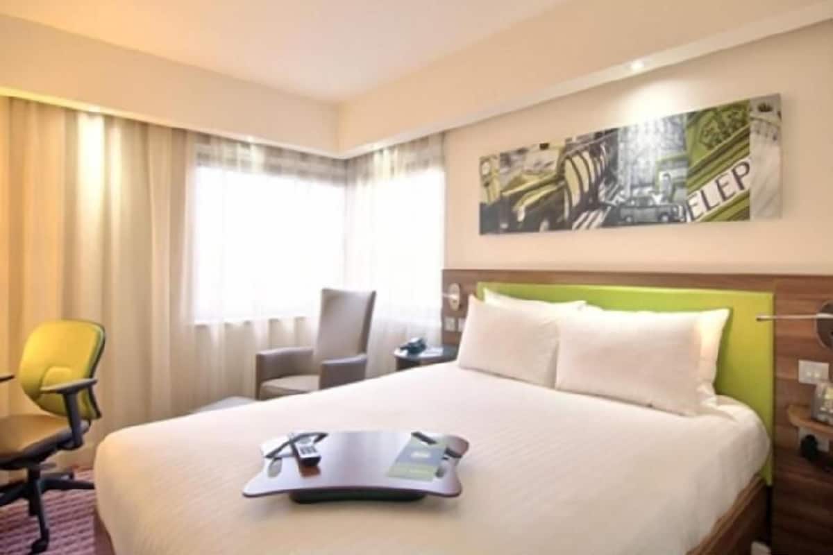 A hotel room with a bed, a chair, a desk, and a painting on the wall.