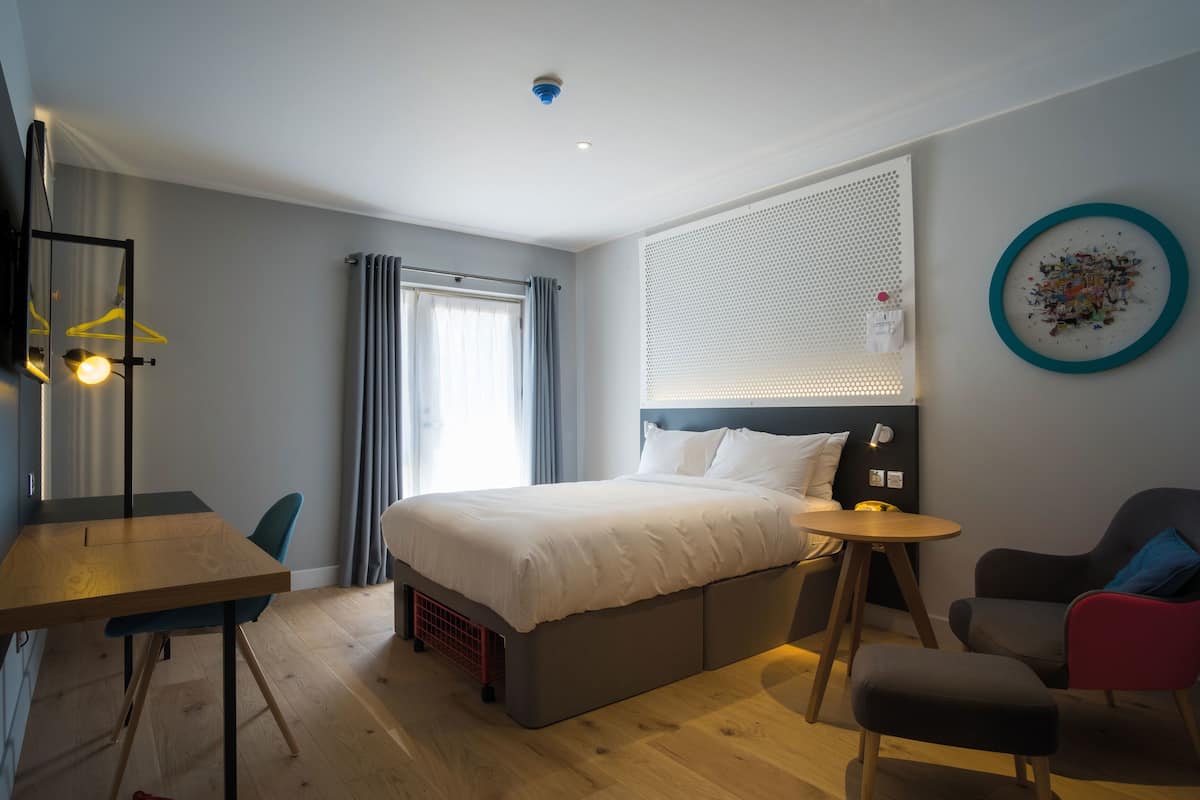 Quarto de hotel moderno com cama, mesa de cabeceira, cadeira e uma pequena mesa redonda.