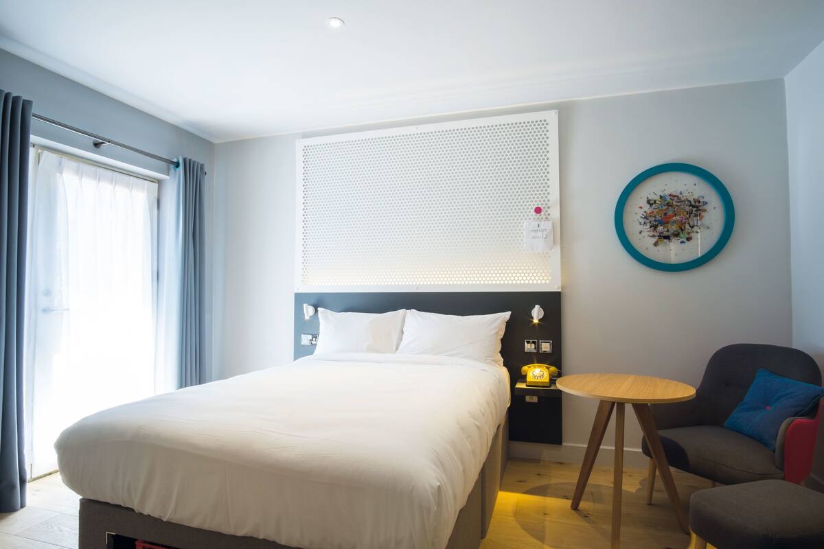 Quarto de hotel com uma cama grande, uma mesa redonda, uma cadeira, uma luminária na parede e um prato decorativo.