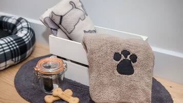 A pet bed, a towel with a paw print, a bone-shaped treat, and a jar with a lid on a dark mat.