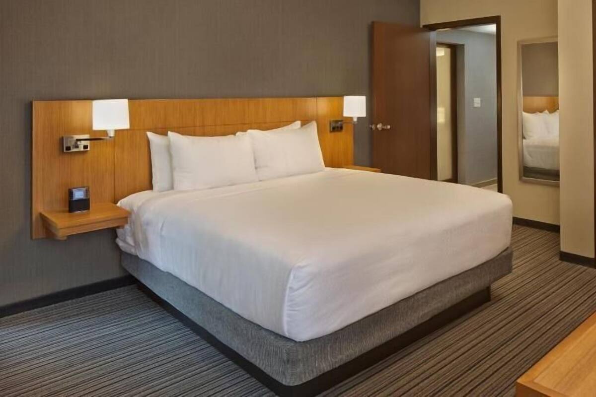 Ein Hotelzimmer mit einem großen Bett, Nachttischlampen, einem Nachttisch, einem hölzernen Kopfteil, einem Teppichboden und einer Tür zu einem weiteren Raum.