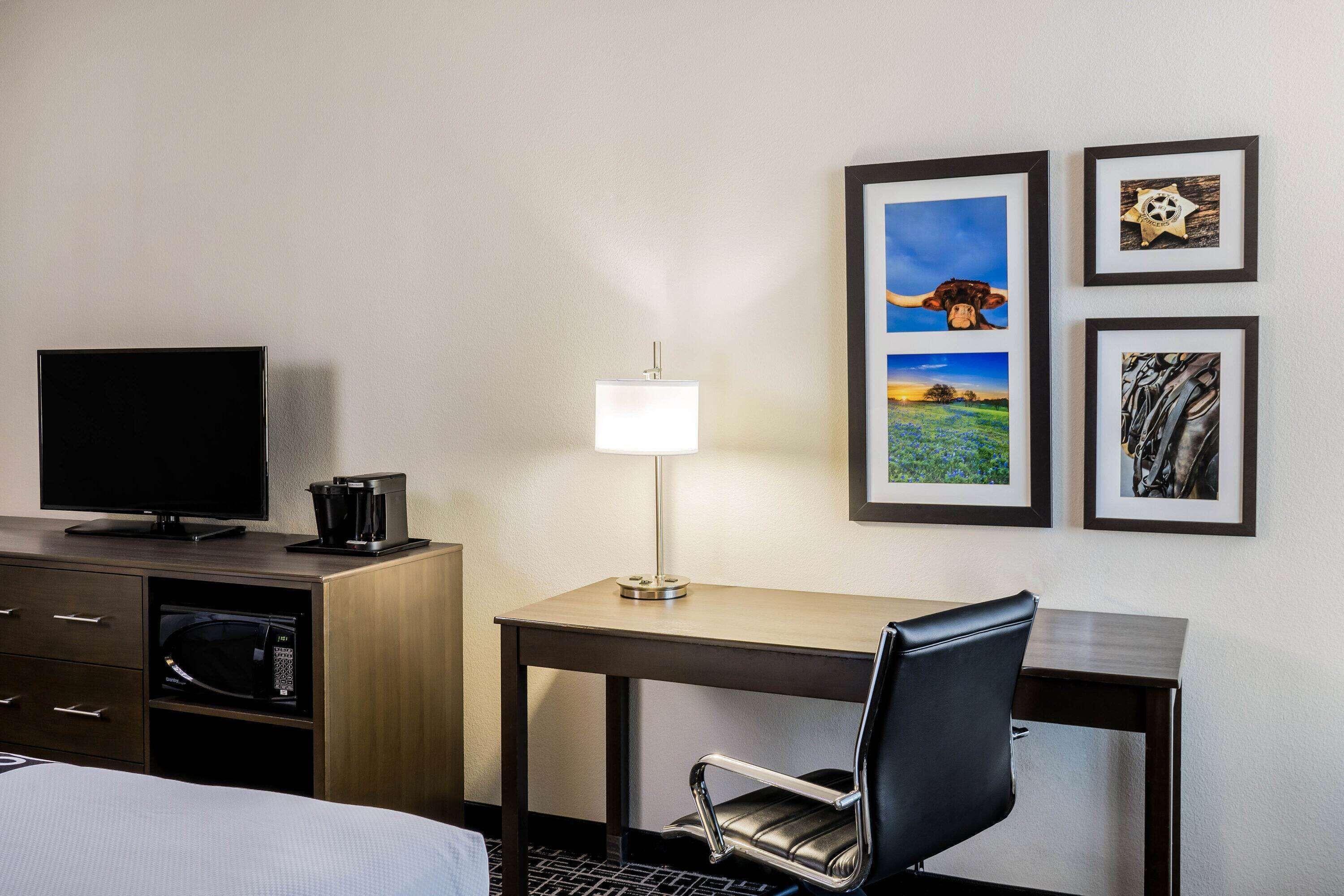 Une chambre d’hôtel avec un lit, un bureau avec une lampe, une télévision, une machine à café, un four à micro-ondes et des tableaux encadrés au mur.