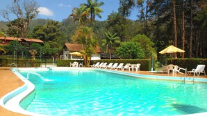 Una piscina con sillas de descanso y sombrillas, rodeada de árboles y un edificio con tejado de baldosas.