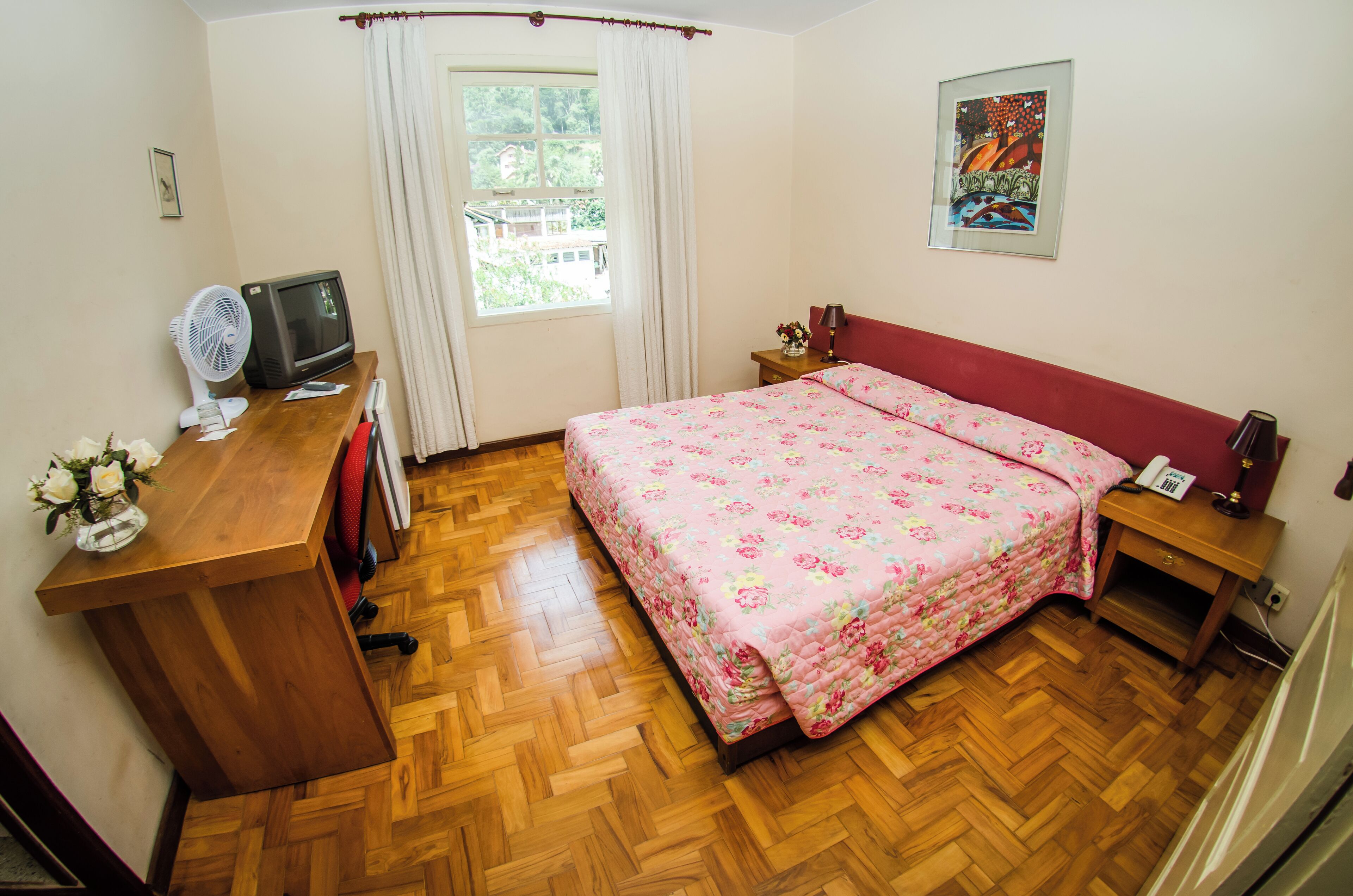 Une chambre avec un plancher en bois, un lit avec une literie à motifs floraux, un bureau avec un ventilateur, une télévision et une fenêtre avec des rideaux.