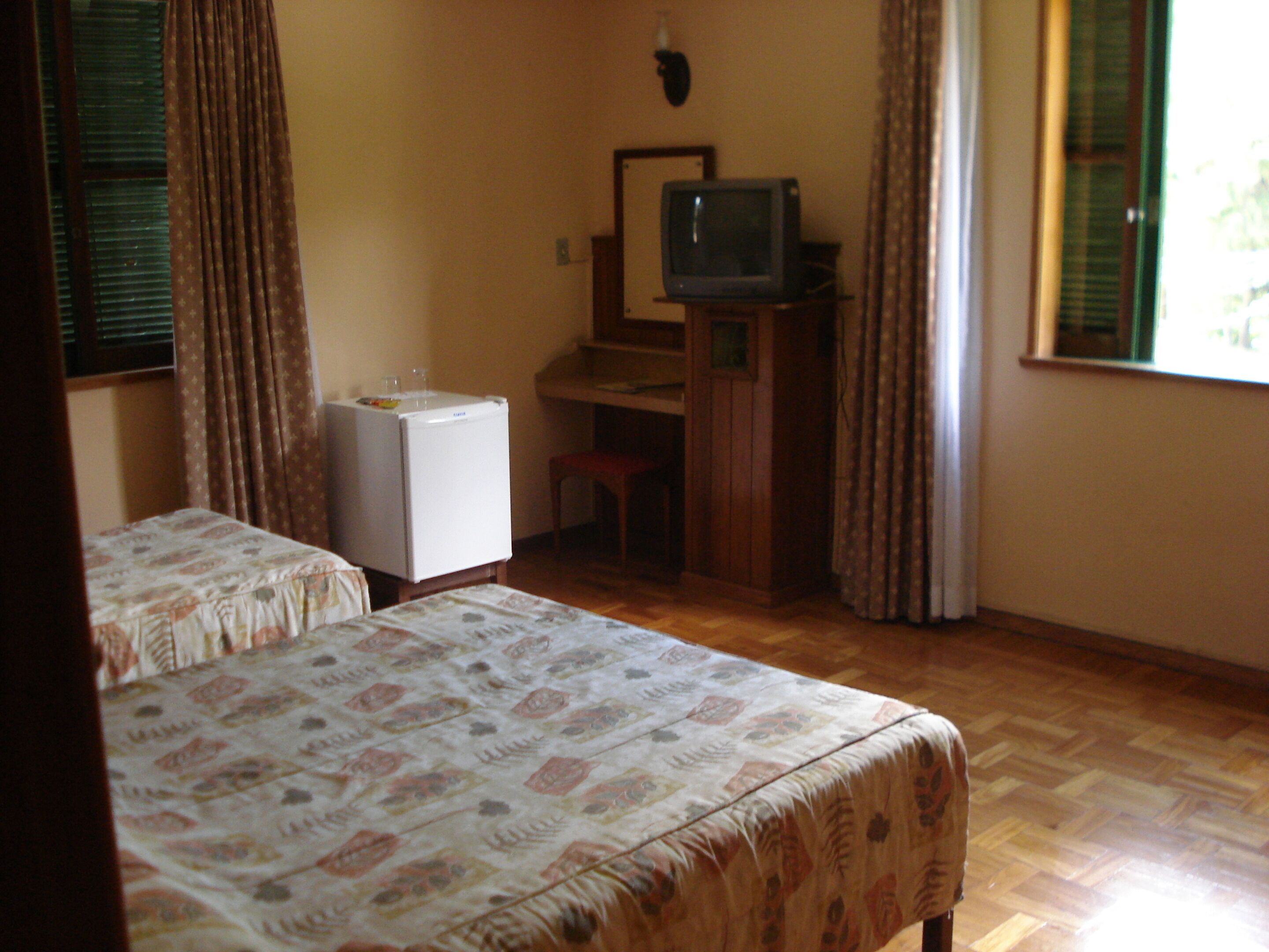 Una habitación con dos camas, un escritorio pequeño con televisor, un refrigerador y una ventana con cortinas.