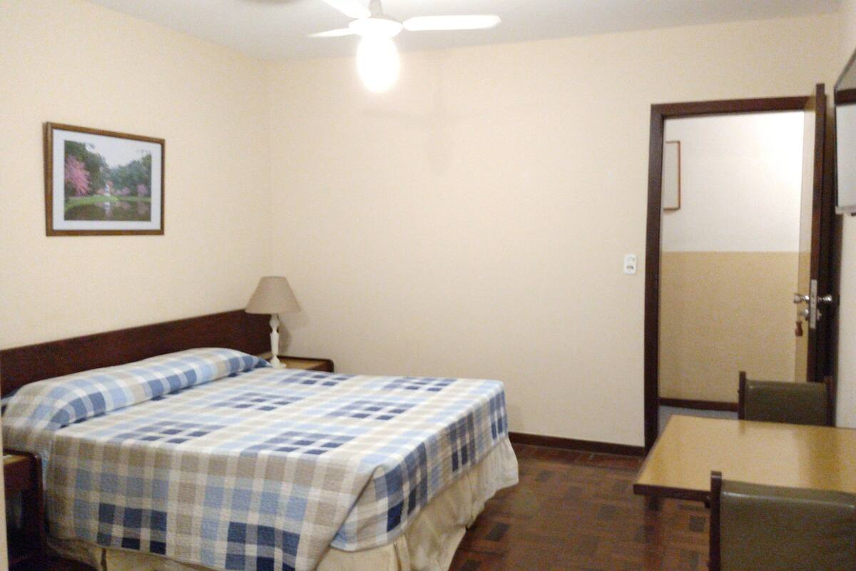 A bedroom with a bed, a table, a chair, a lamp, a TV, and a door.