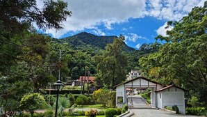 Un camino de adoquines que conduce a un edificio blanco con techo marrón, rodeado de abundante vegetación y una montaña al fondo.