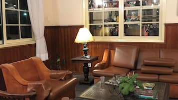 A living room with a brown leather sofa, a wooden coffee table, and a lamp. There is a window with a view of a building and a shelf with various items.