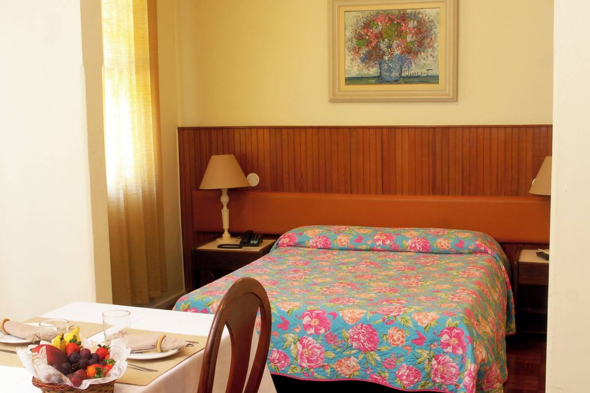 A hotel room with a bed, a table with a basket of fruit, a chair, a nightstand with a lamp, and a painting on the wall.