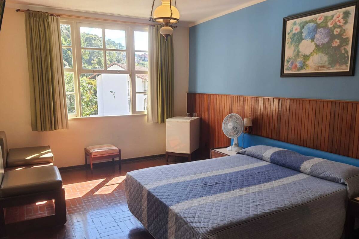 A hotel room with a bed, a chair, a small table, a standing fan, and a window with curtains.