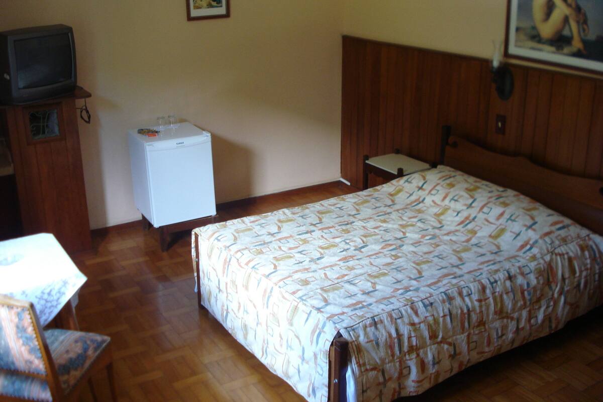 A bedroom with a bed, a small white refrigerator, a television on a wooden cabinet, and a chair.