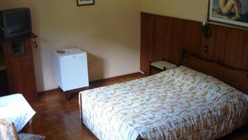 A bedroom with a bed, a small white refrigerator, a television on a wooden cabinet, and a chair.