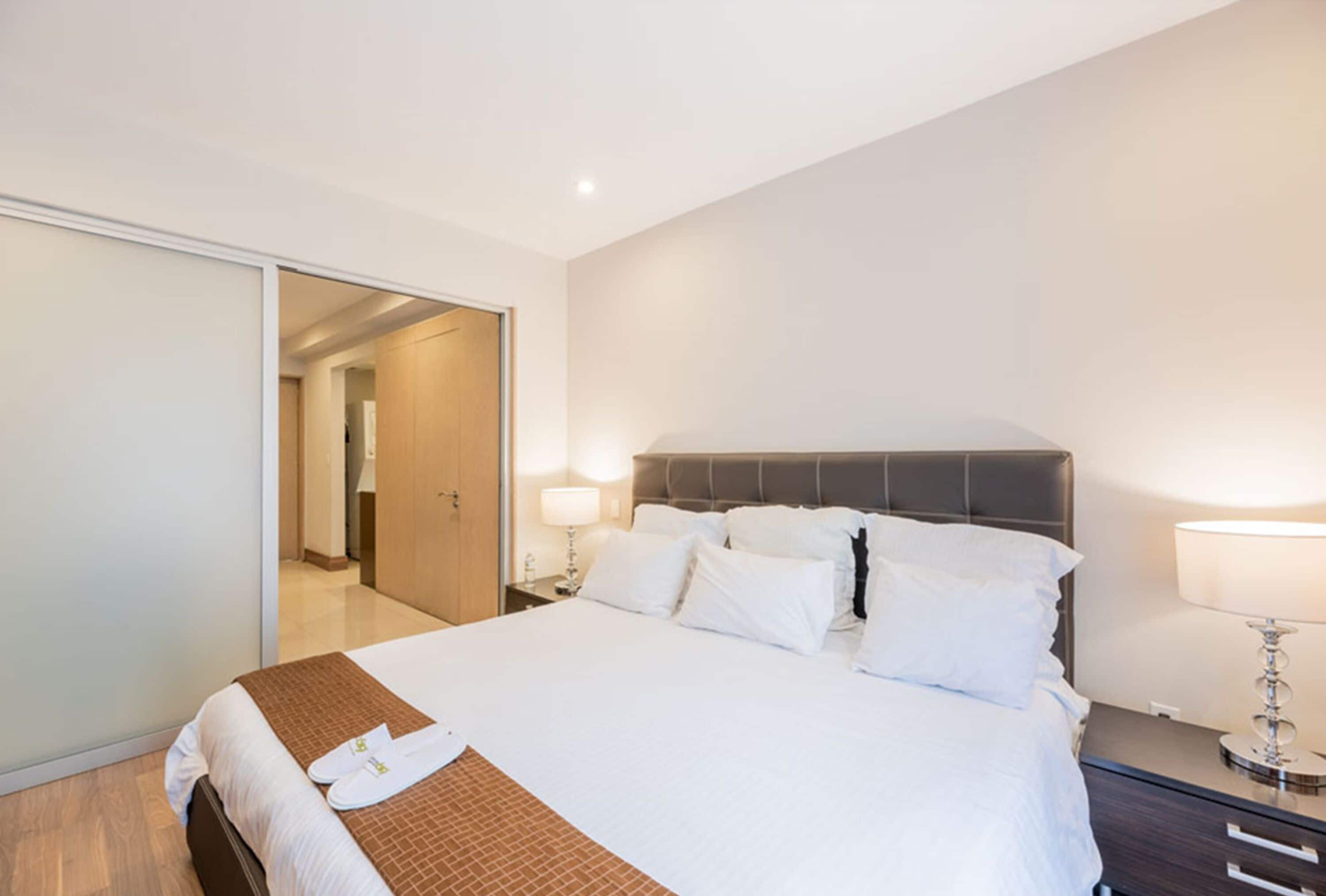 Habitación de hotel con una cama grande, piso de madera, armario con puerta corredera y mesita de noche con lámpara.