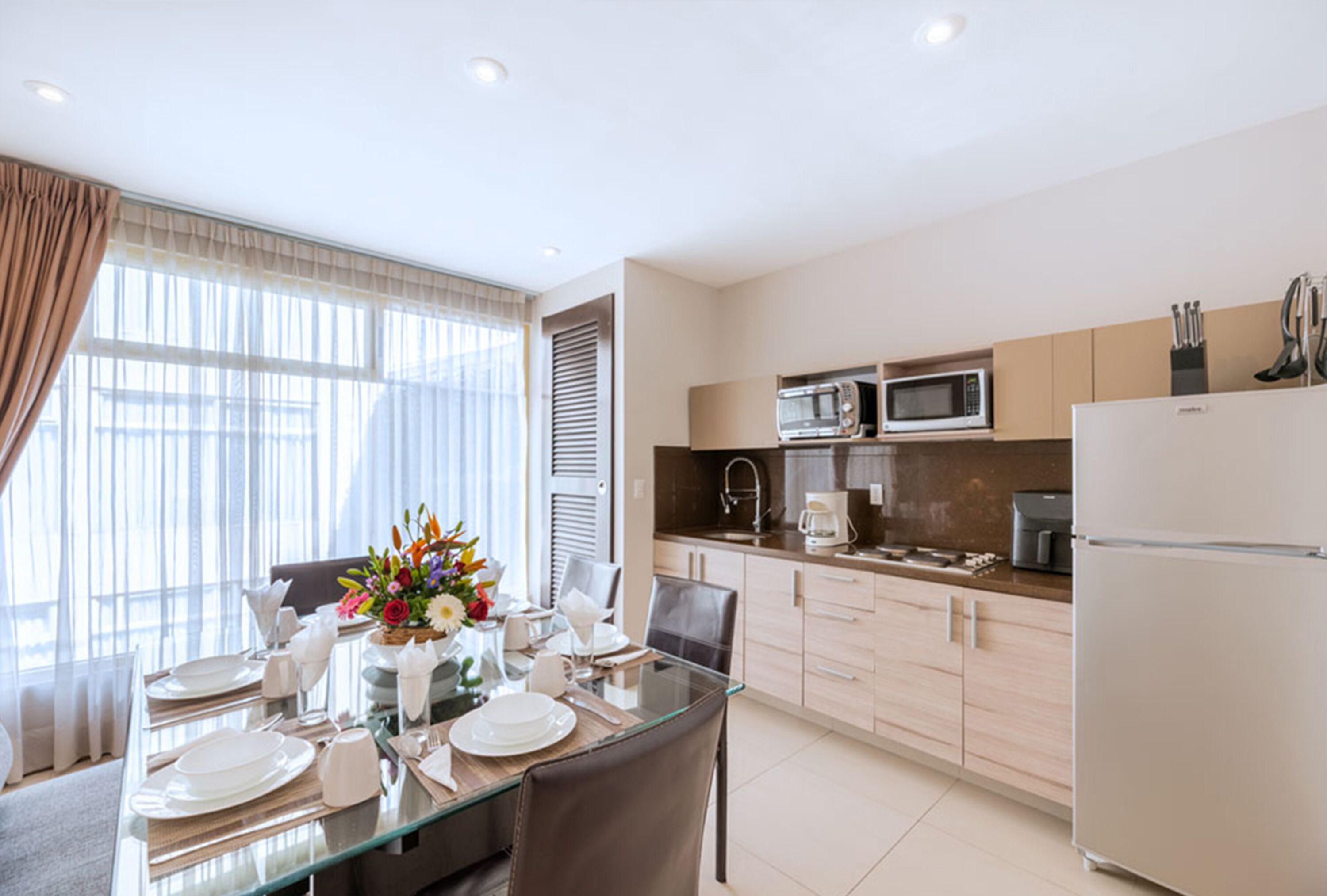 Una cocina moderna con comedor, que incluye una mesa de vidrio, electrodomésticos de acero inoxidable y un ventanal con cortinas.