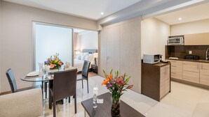 Una habitación de hotel moderna con zona de comedor, kitchenette y baño visible a través de una puerta de cristal.