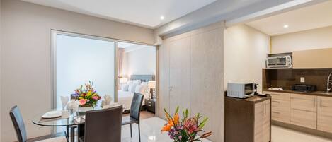 A modern hotel room with a dining area, kitchenette, and a bathroom visible through a glass door.