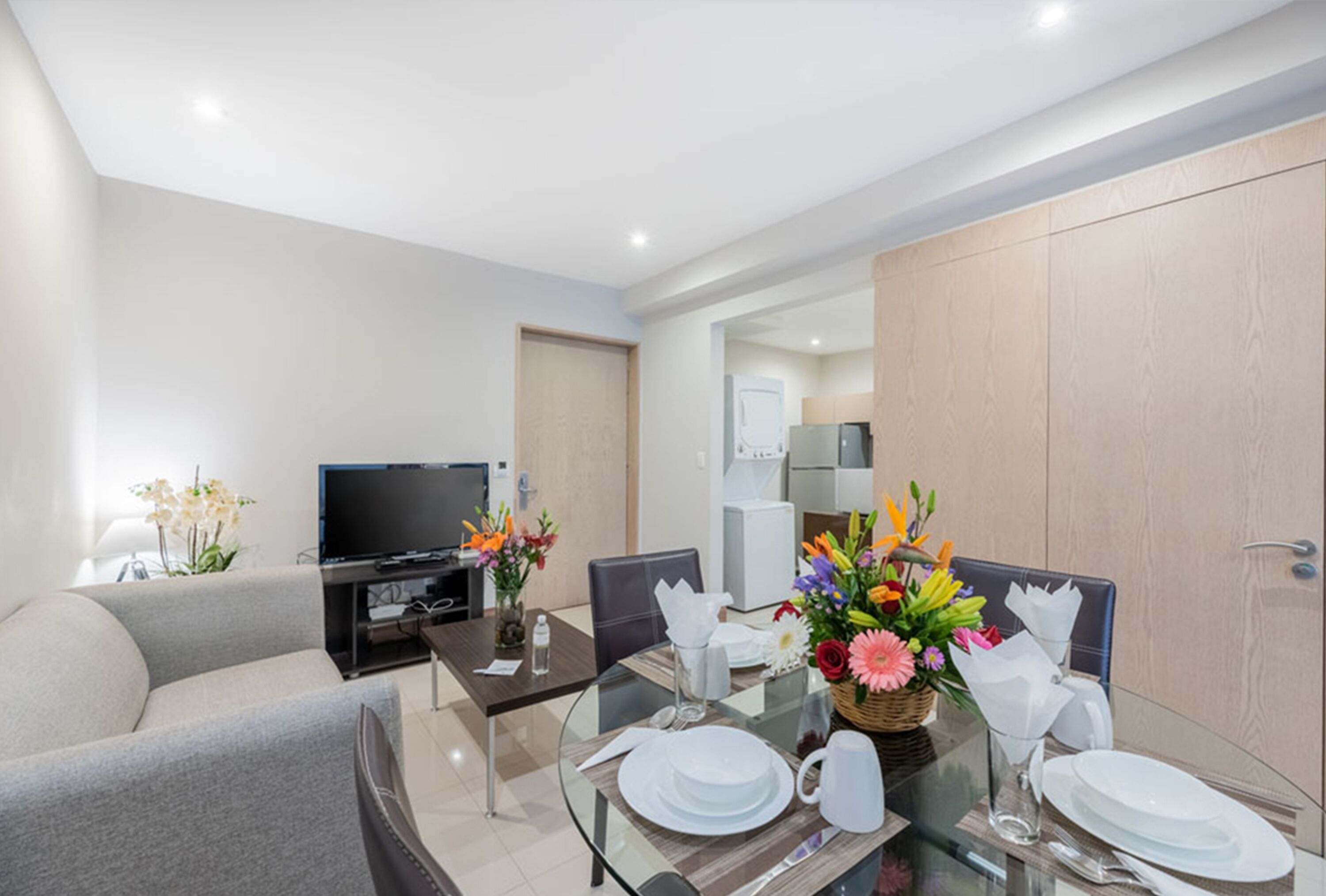 A modern hotel room with a dining area, a sofa, a TV, and a kitchen in the background.