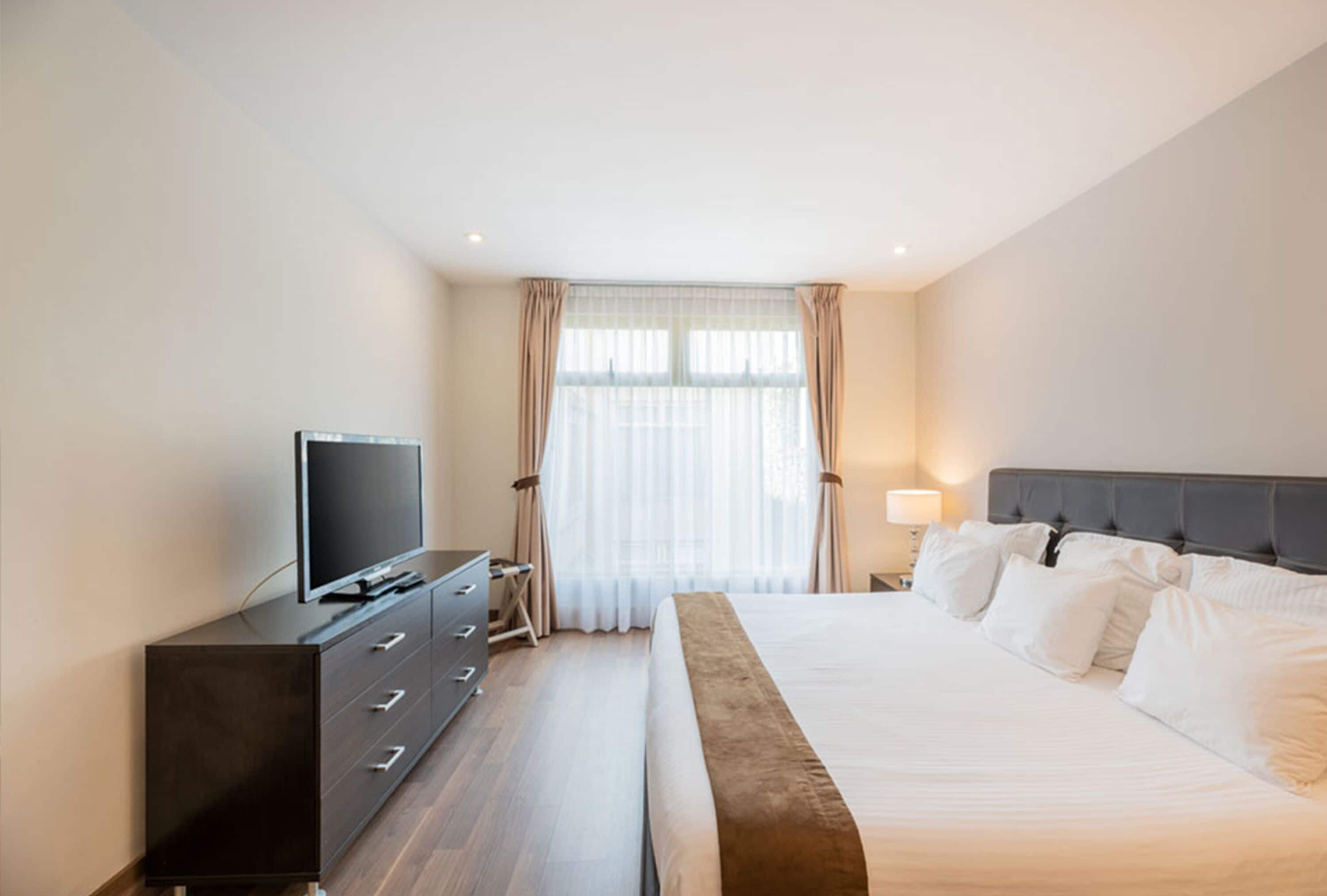 Una habitación de hotel moderna con una cama grande, un televisor de pantalla plana sobre una cómoda y una ventana con cortinas translúcidas.
