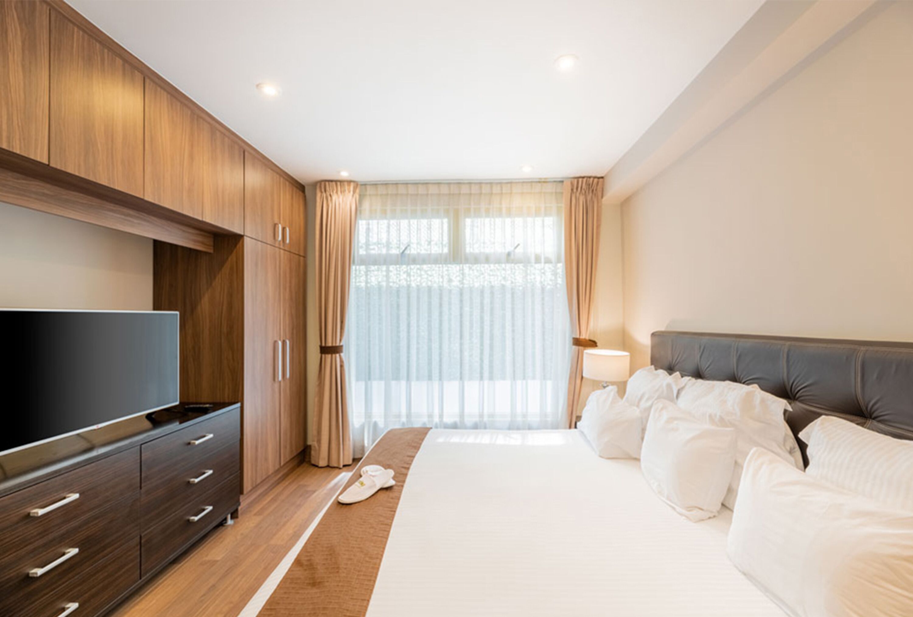 Ein modernes Hotelzimmer mit einem großen Bett, einem Flachbildfernseher, Holzschränken und einem großen Fenster mit durchscheinenden Vorhängen.