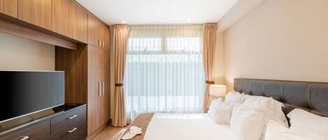 Ein modernes Hotelzimmer mit einem großen Bett, einem Flachbildfernseher, Holzschränken und einem großen Fenster mit durchscheinenden Vorhängen.