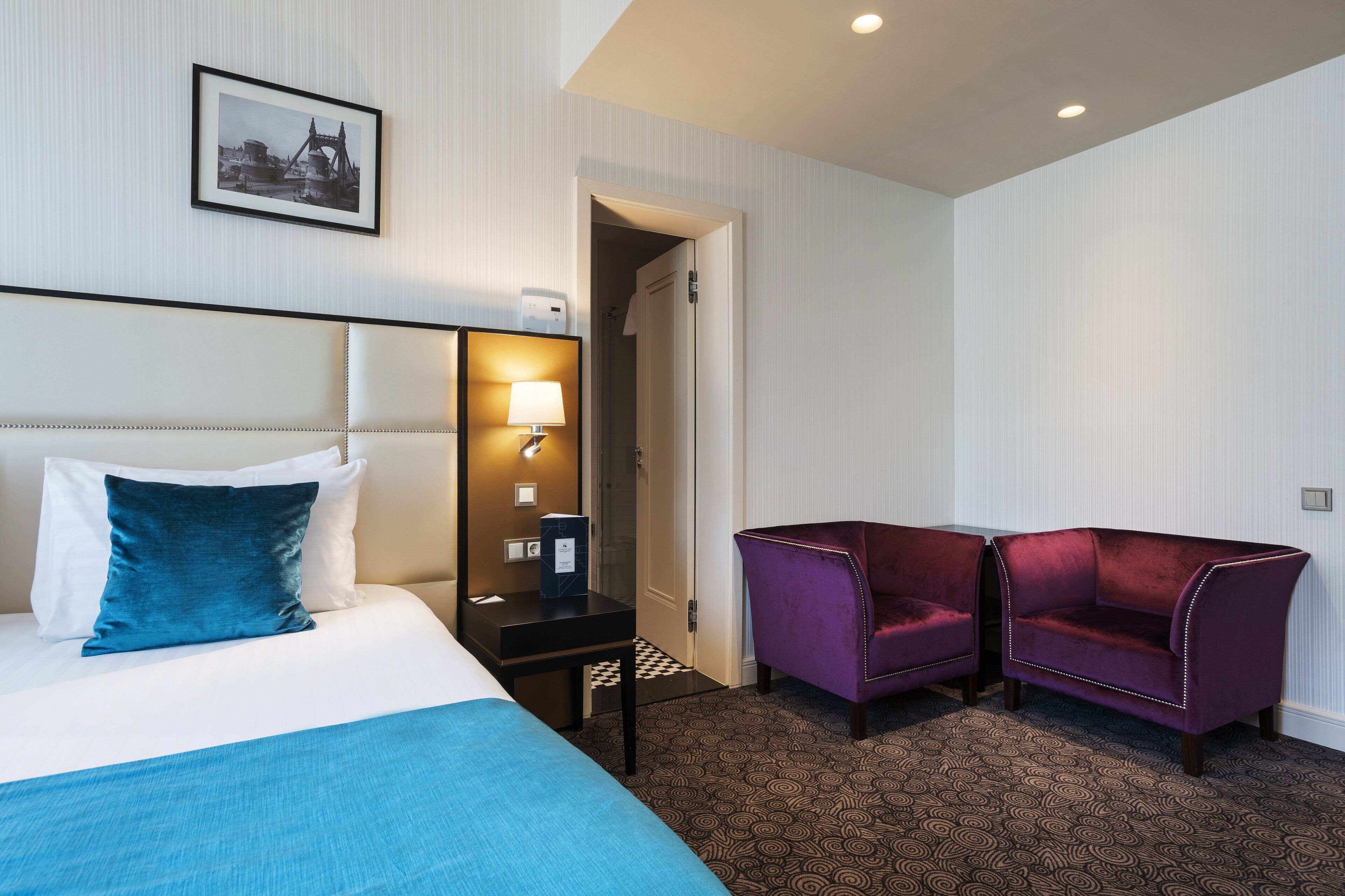 A hotel room with a bed, a nightstand with a lamp, two purple armchairs, and a patterned carpet.