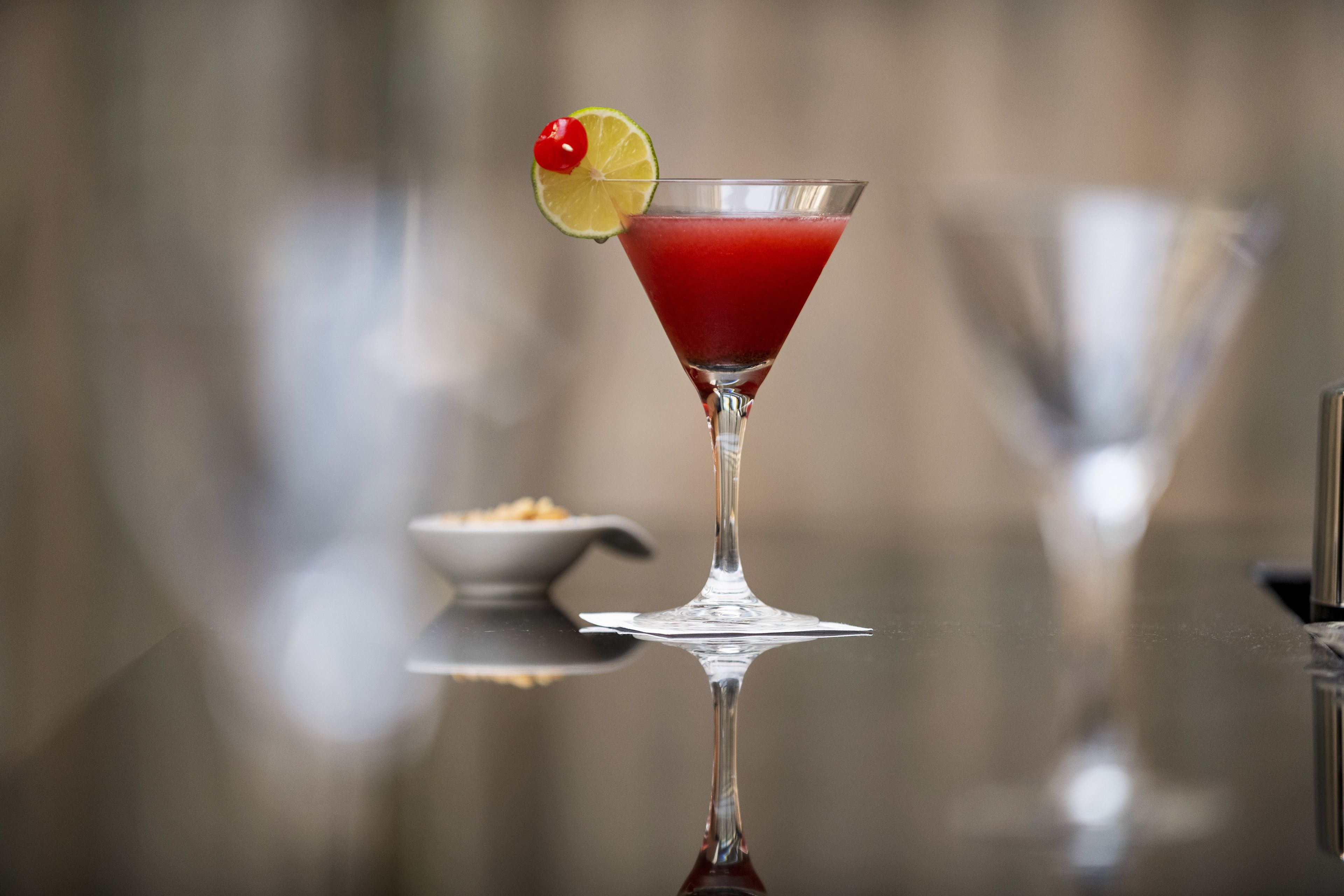 A martini glass with a red drink, garnished with a cherry and a slice of lime, on a reflective surface.