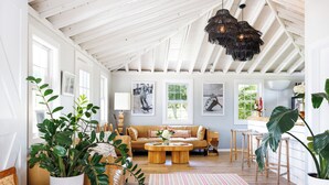 A spacious living room with a high ceiling, wooden beams, a large sofa, a coffee table, and a dining area with stools. - Hero Beach Club (Montauk)