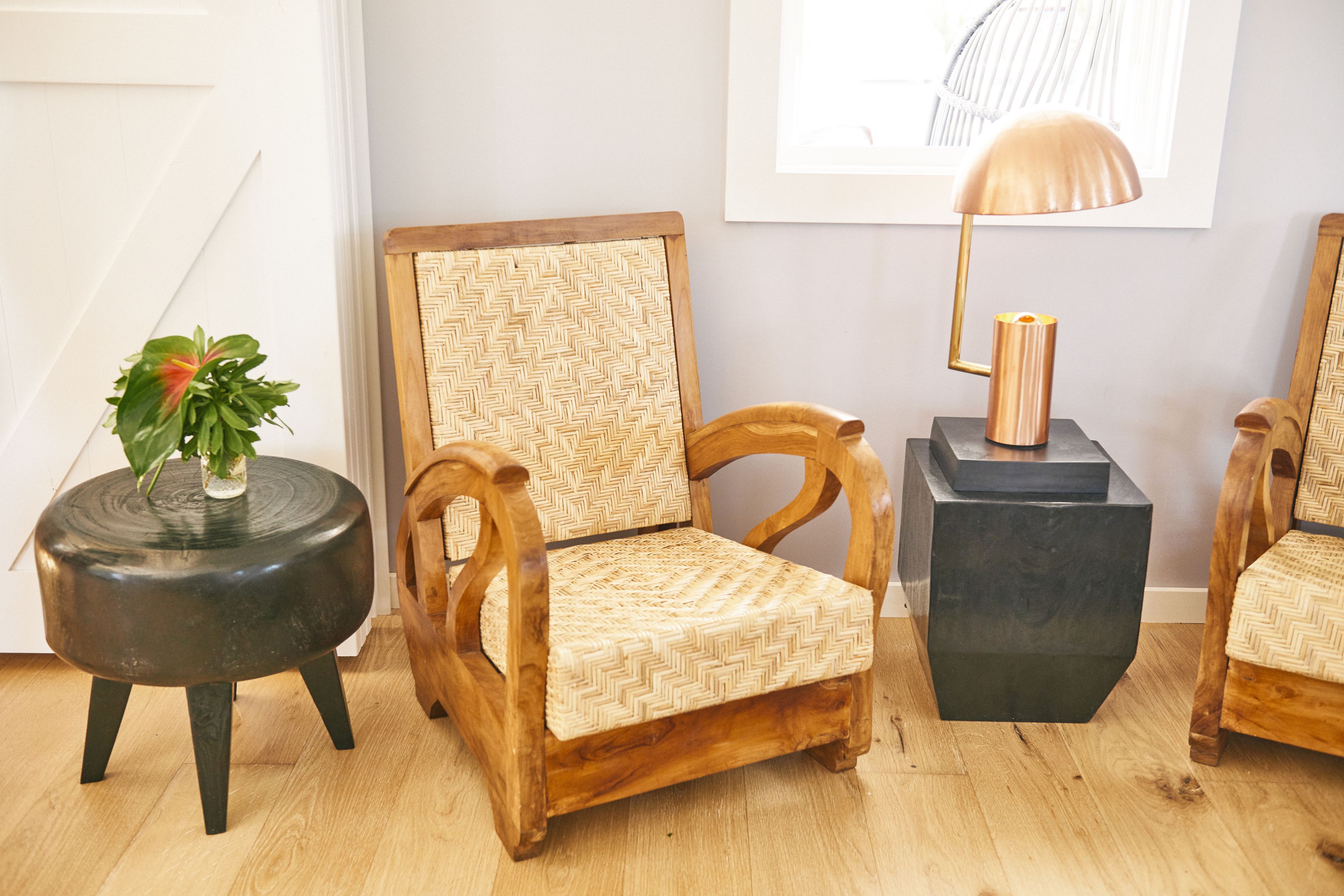 A wooden chair with a woven backrest, a round black side table with a plant, and a black cube-shaped side table with a lamp.