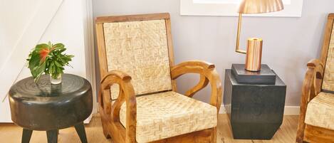 A wooden chair with a woven backrest, a round black side table with a plant, and a black cube-shaped side table with a lamp.