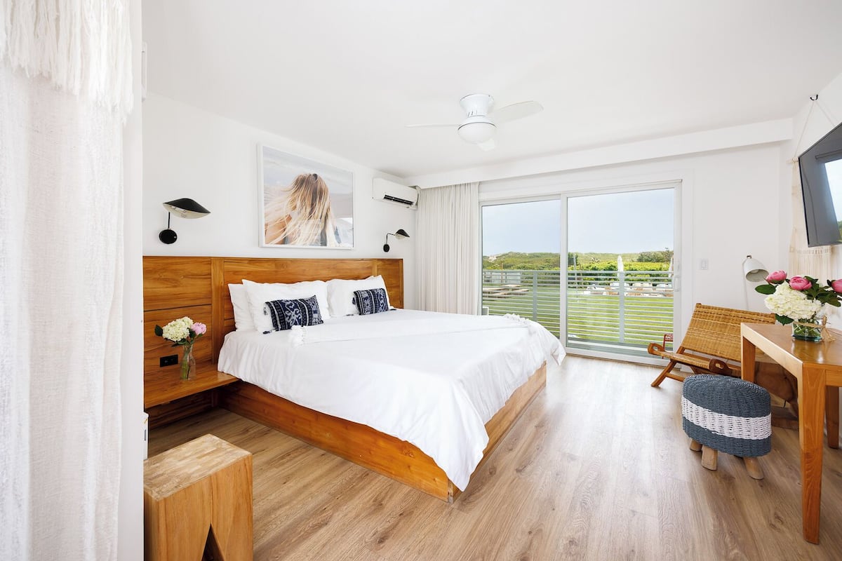 A bedroom with a large bed, a wooden headboard, a nightstand, a desk with a chair, a television, and a balcony with a view.