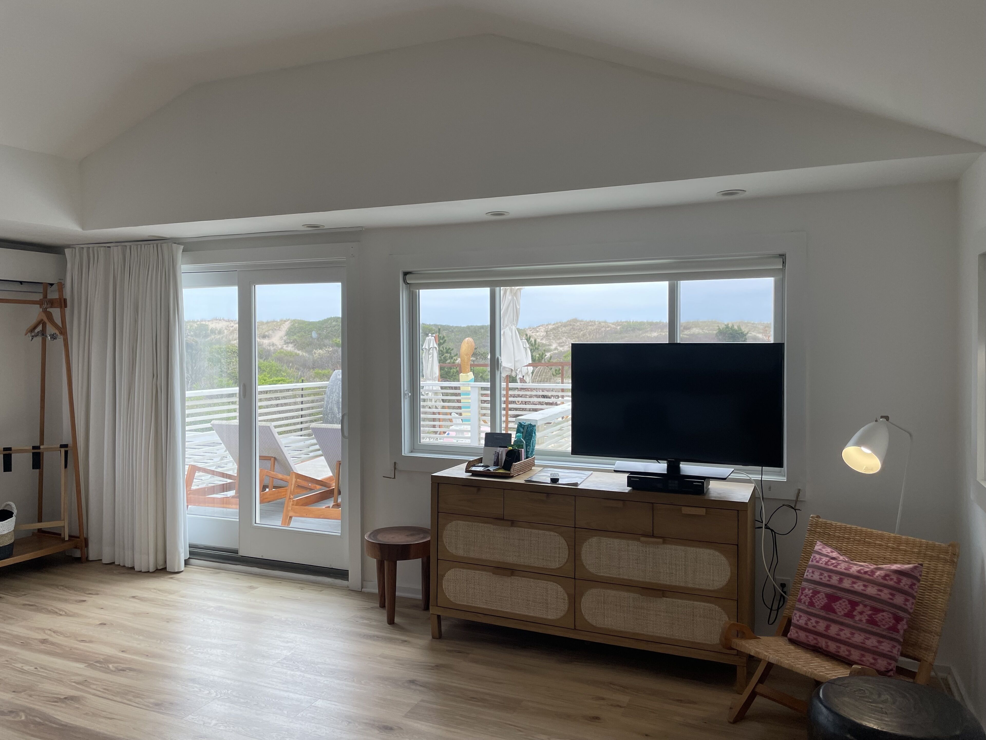 Ein geräumiges Zimmer mit großem Fenster, einem Flachbildfernseher, einem Holzkasten, einem Korbstuhl und einem kleinen Hocker.