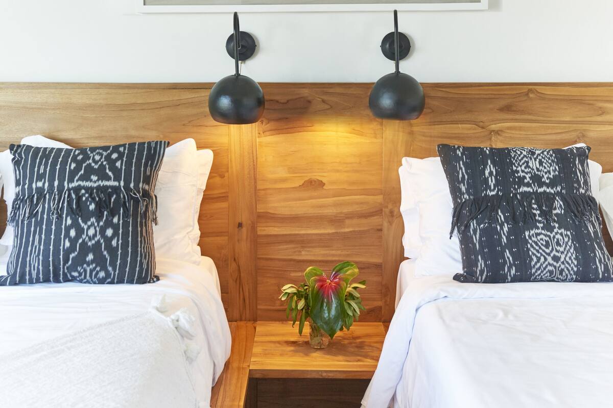 A room with two beds, a wooden headboard, a small table with a plant, and a framed picture above the beds.
