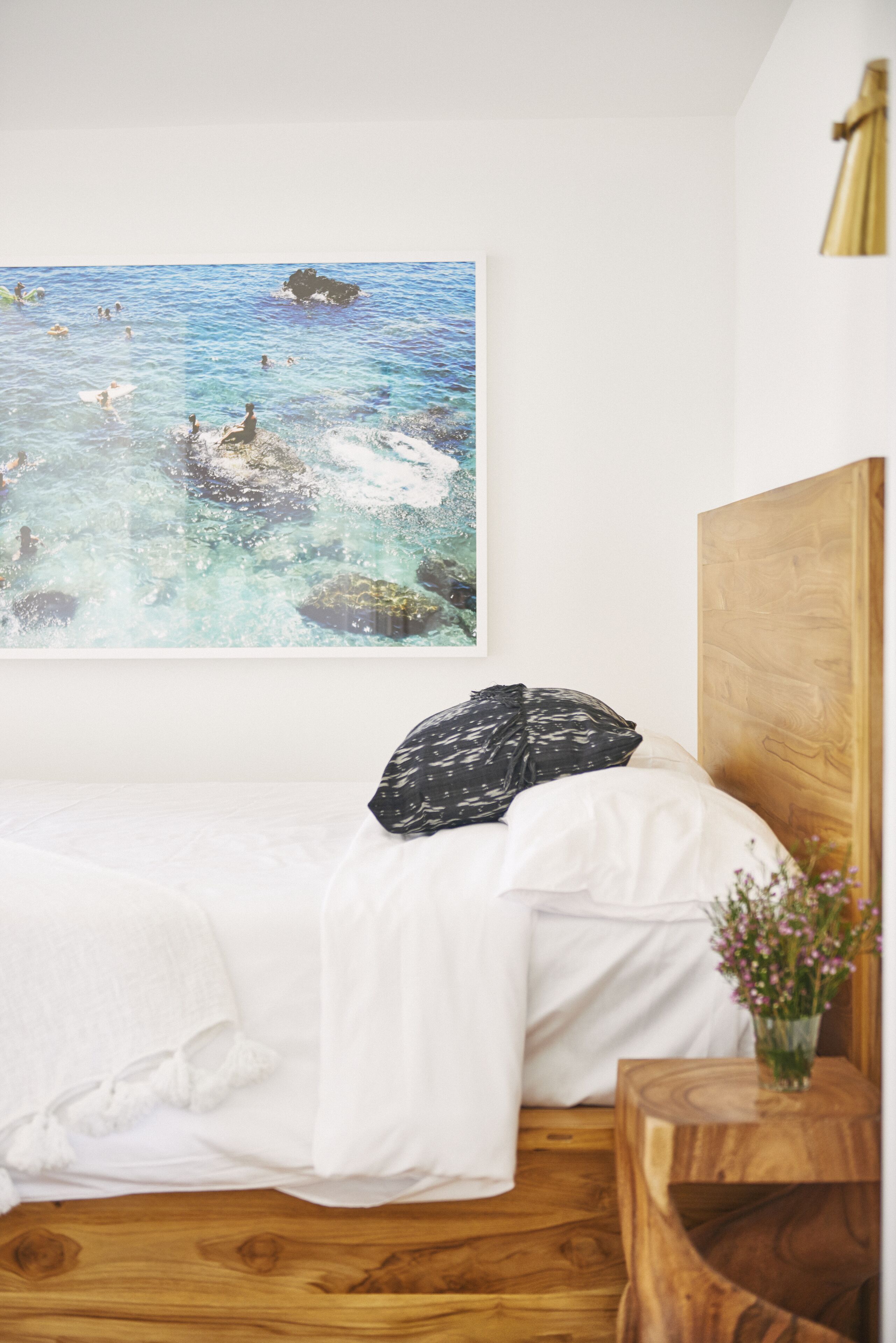 Un dormitorio con cama, cabecera de madera, cuadro colgado en la pared que muestra una playa y una mesita de noche de madera con un florero.