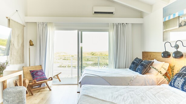 A bedroom with two beds, a wooden rocking chair, a small table, and a view of the outdoors. - Hero Beach Club (Montauk)