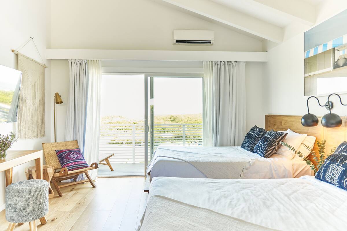 A bedroom with two beds, a wooden rocking chair, a small table, and a view of the outdoors.