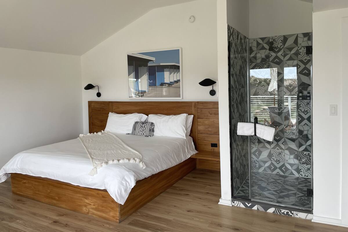 A modern bedroom with a wooden bed, a ceiling fan, a large mirror, and a shower with patterned tiles.